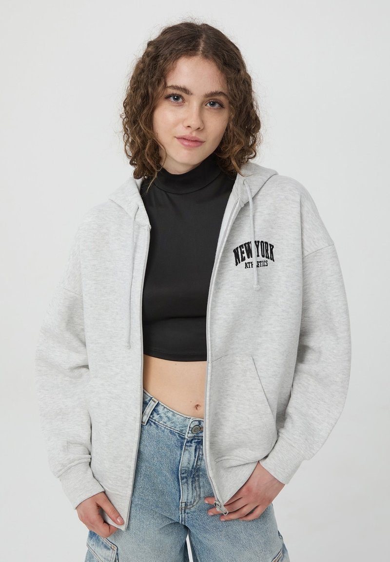Felpa grigia con zip e tasca frontale, con testo "NEW YORK" in nero. Indossata sopra un top nero corto, accompagnata da un paio di pantaloni corti in denim larghi.