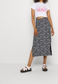 Mujer con sandalias blancas, falda midi negra con un patrón floral blanco y una camiseta corta blanca con un gráfico de pizza rosa "WAP".