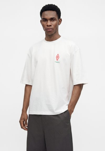 Νέος άνδρας φοράει άνετο λευκό T-shirt με μικρό κόκκινο γεωμετρικό λογότυπο και κείμενο, σε συνδυασμό με σκούρες γκρι βερμούδες σε απλό φόντο.