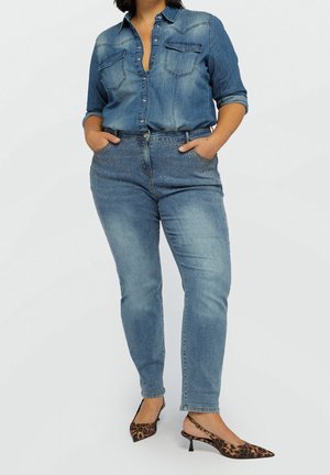 Donna che indossa una camicia di jeans con bottoni e jeans skinny borchiati con tacchi slingback leopardati, in piedi con le mani in tasca.