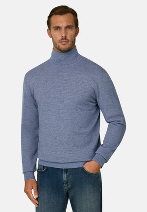 Homme portant un pull à col roulé bleu clair et un jean bleu, debout avec une main dans la poche devant un fond uni.