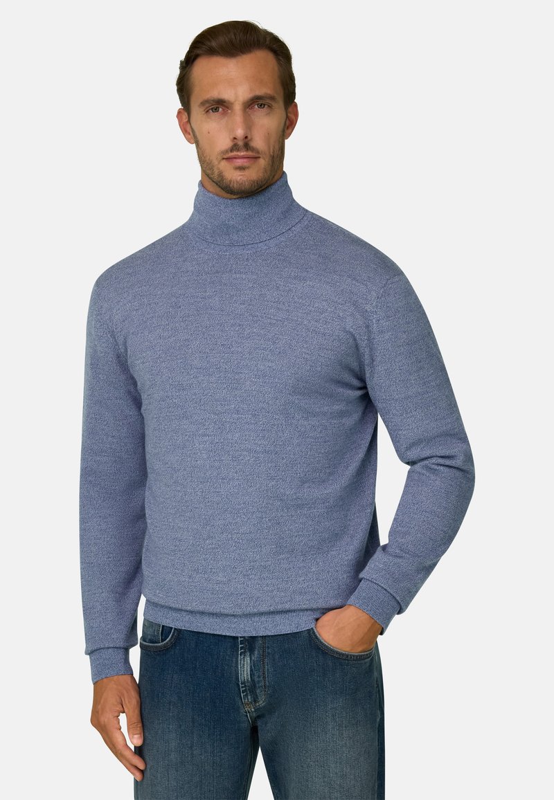 Homme portant un pull à col roulé bleu clair et un jean bleu, debout avec une main dans la poche devant un fond uni.