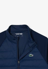 Giacca leggere di colore blu navy con zip frontale, sezioni imbottite e logo di coccodrillo verde sul petto. Design con colletto.