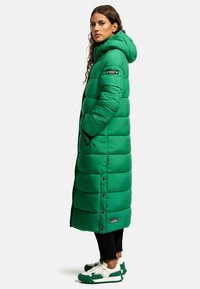 Manteau long matelassé vert avec capuche, présentant un design matelassé, des poches latérales, des boutons-pression et un patch logo sur la manche.