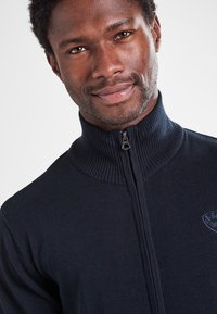 Pull en maille bleu marine avec un col montant zippé, une texture côtelée et un petit logo sur le côté gauche de la poitrine. Finition du tissu lisse et coupe ajustée.