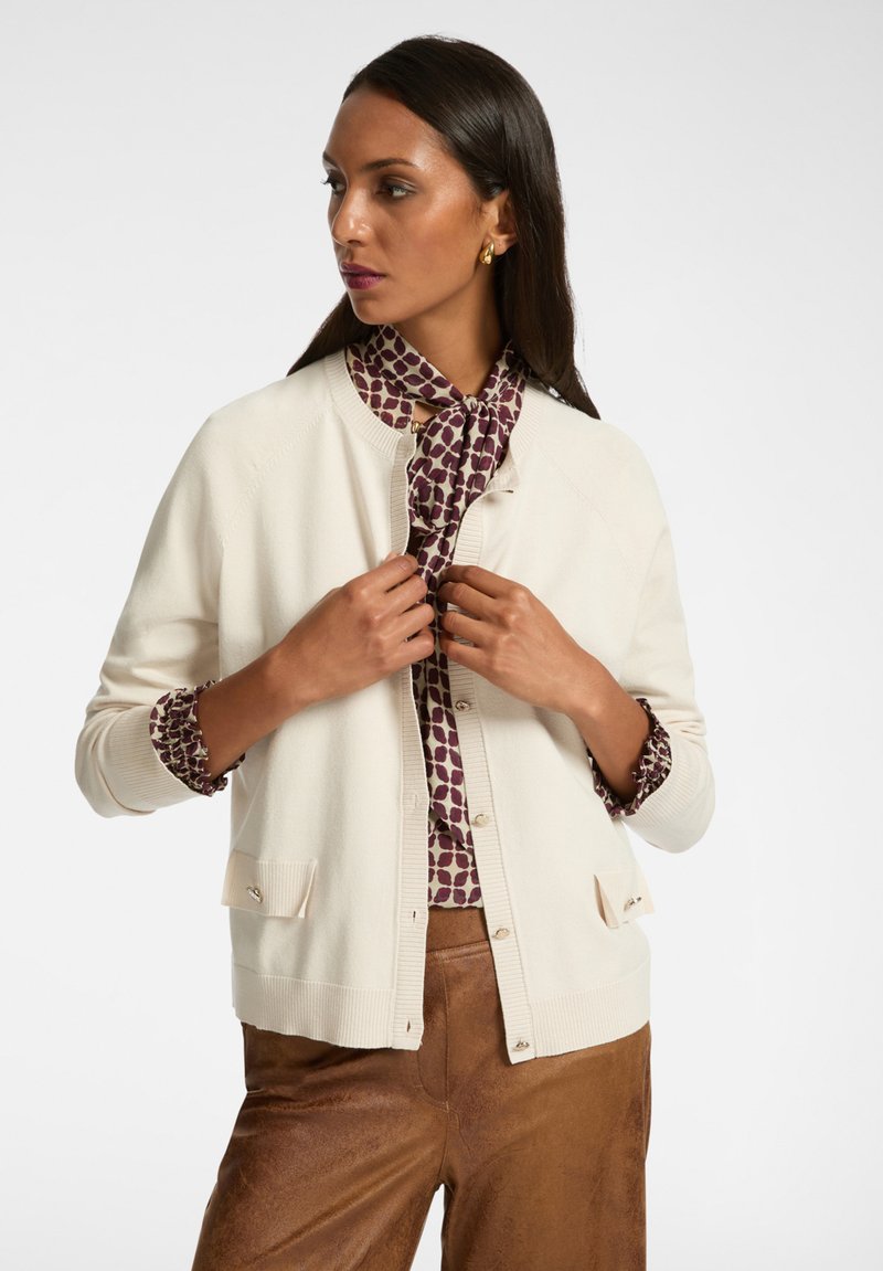 Cremfarbener Cardigan mit geripptem Saum und zwei vorderen Taschen. Darunter eine kastanienbraune, gemusterte Bluse mit einem gebundenen Halstuch.