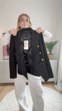 Blazer preto texturizado com três botões dourados e interior em cetim, exibido contra um fundo claro com uma planta em vaso nas proximidades.