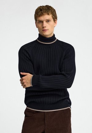Giovane uomo con capelli corti e ricci che indossa un maglione a collo alto nero a coste con bordo beige e pantaloni marroni, in piedi con le braccia incrociate.