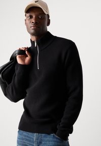 Black ribbed knit sweater with a high collar and half-zip, paired with light blue denim. Model holding a black bag. Beige cap accent.
