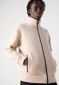 Beige fleece jacket with a high collar, front zipper, and side pockets. Features a textured surface and a relaxed fit.