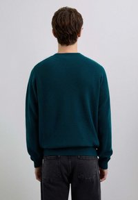 Suéter de punto color verde azulado con cuello redondo, puños y dobladillo acanalados. Llevado sobre unos vaqueros gris oscuro, visto desde atrás.