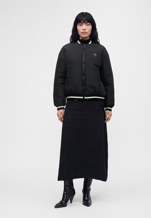 Femme aux longs cheveux noirs portant une veste bomber noire, une jupe midi noire et des bottines noires à talons, debout devant un fond uni.