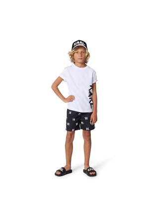 T-shirt blanc avec un détail graphique noir, short noir avec des motifs de logo blancs, et sandales noires, portés par un enfant aux cheveux bouclés.