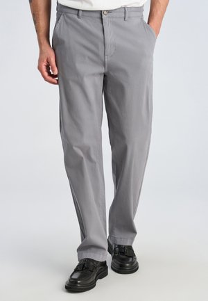 Man draagt lichtgrijze recht gesneden broek gecombineerd met zwarte tassel loafers, staat met één hand in de zak.