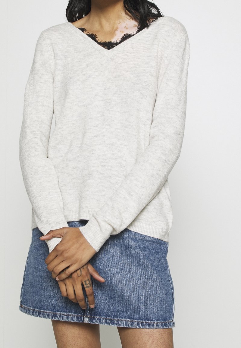 Jasnoszary sweter dzianinowy z dekoltem w kształcie V, zestawiony z dżinsową mini spódnicą. Posiada długie rękawy i luźny krój.