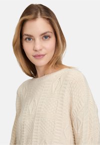 Helle beigefarbene Strickjacke mit strukturiertem Muster und erhobenen geometrischen Designs sowie einem weiten Ausschnitt. Weiches Material mit langen Ärmeln.