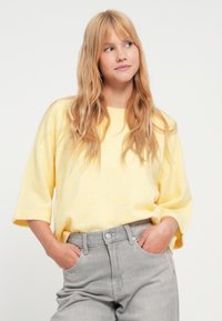 Dzeltena vaļīga džemperis ar trīs ceturtdaļu piedurknēm, izgatavots no mīksta, fakturēta auduma; kombinēts ar gaiši pelēkajām augsta vidukļa džinsu biksēm.