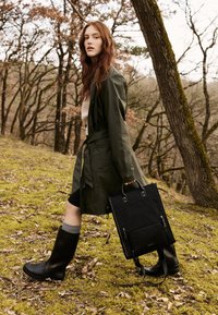 Black canvas tote bag with dual handles and zippered pockets, paired with a green waterproof coat, black rubber boots, and a textured sweater.
