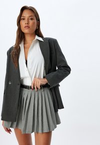 Blazer grigio sopra una camicia bianca, abbinato a una gonna a pieghe grigia a scacchi. Il blazer ha rever a punta e due bottoni.