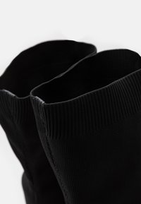 Detailní záběr na vroubkované, elastické horní okraje páru černých látkových šněrovacích bot na jednobarevném světlém pozadí.