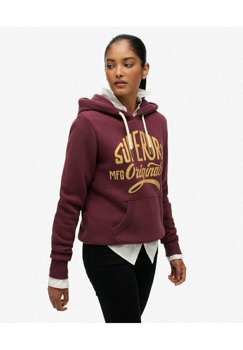 Bordowy hoodie z dużym żółtym logo "SUPERDRY", z trokami, kieszenią z przodu i ściągaczami, nałożony na białą koszulę z kołnierzykiem.