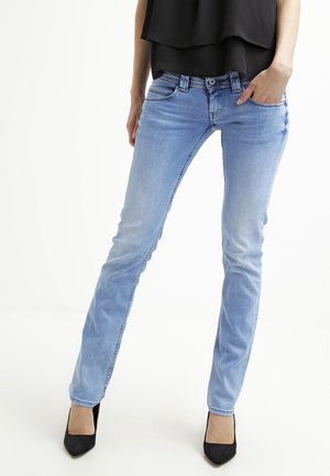 Mujer con jeans ajustados azul claro y tacones altos negros, de pie con una mano en el bolsillo frente a un fondo blanco liso.