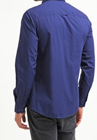 Marinblå långärmad skjorta med knäppkrage, texturerad tyg, axelsömmar och uppvikta ärmar, visad bakifrån.