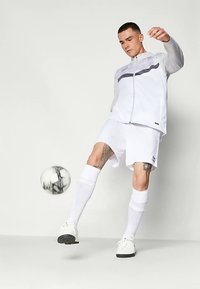 Hombre vestido con ropa deportiva blanca y calcetines controlando un balón de fútbol en el aire, con un fondo sencillo y tatuajes visibles en sus piernas.