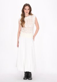 Leichtes, cremefarbenes Strickoberteil mit einem Spitzenmuster, kombiniert mit einem weißen Maxirock und schwarzen Stiefeletten. Das Outfit hat eine lockere Passform.