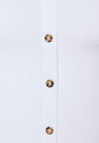 Vêtement blanc à boutons, ornée de cinq boutons ronds, brillants et de couleur dorée-brun à l'avant, avec une texture de tissu lisse.