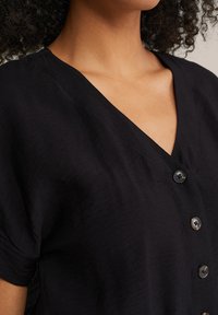 Čierny top s krátkym rukávom a hlbokým výstrihom do V, ktorý má zapínanie na gombíky s čiernymi gombíkmi a mäkkou štruktúrovanou tkaninou.