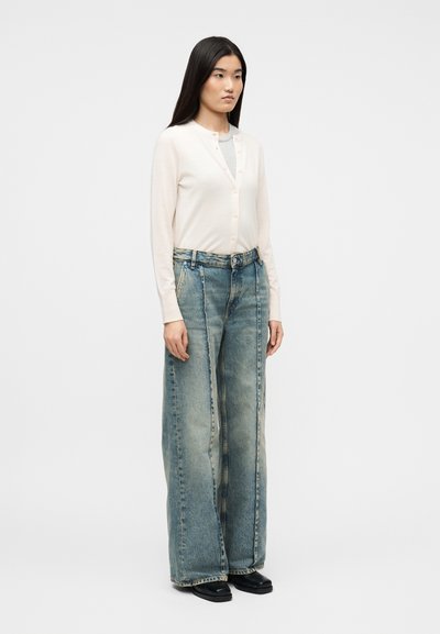 Γυναίκα όρθια, φορώντας ένα κρεμ ζακετάκι με κουμπιά πάνω από γκρίζο τοπ, φαρδιά ξεβαμμένα τζιν παντελόνια και μαύρα παπούτσια, μπροστά σε απλό ανοιχτό φόντο.