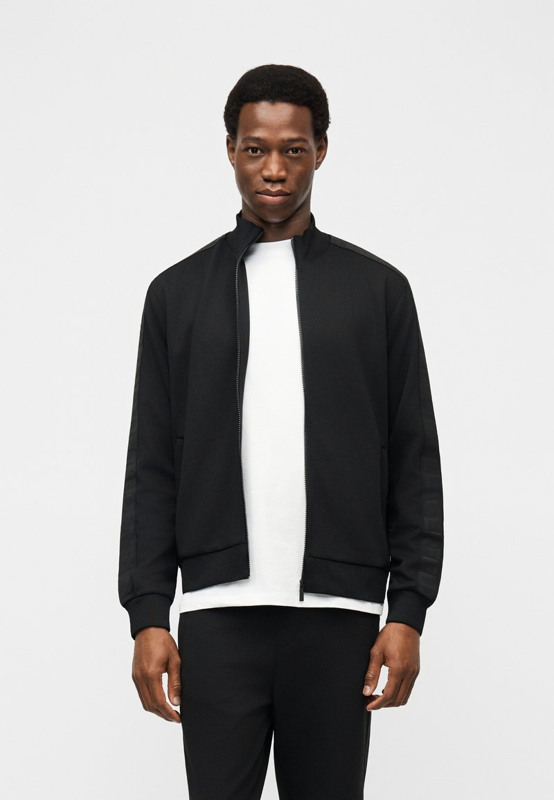 Νέος άνδρας με μαύρο φούτερ με φερμουάρ πάνω από λευκό πουκάμισο, στέκεται μπροστά σε απλό ανοιχτό γκρι φόντο, κοιτάζοντας προς τα εμπρός.