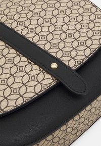 Borsa in tessuto beige e nera con un motivo geometrico composto da cerchi e quadrati alternati, fissata con una tracolla nera con un bottone a scatto dorato.