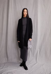 Schwarzer langer Cardigan über einem dunkelgrauen gerippten Top und einem grauen Wollrock. Hält eine hellgraue Strick-Clutch. Schwarze Stiefeletten vervollständigen das Outfit.