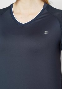 Tmavě šedé sportovní tričko s výstřihem do V, se bílým lemem, vyrobené z hladké tkaniny a s malým reflexním logem na přední straně.