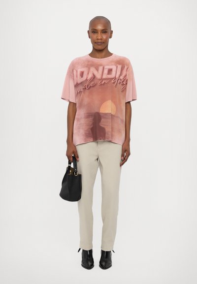 T-shirt rose surdimensionné avec un graphique de coucher de soleil et le texte "Dondi made in Italy". Associé à un pantalon beige et un sac à main noir, porté avec des bottines.