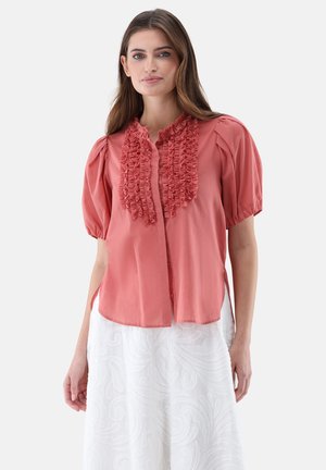 Korallenfarbene Bluse mit Puffärmeln und Rüschen entlang der Vorderseite. Strukturierter Stoff, Knopfverschluss und seitliche Schlitze verleihen dem Design eine besondere Note.