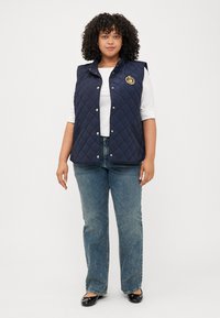 Una donna dai capelli ricci sta in piedi, indossando un gilet trapuntato blu scuro sopra una camicia bianca e jeans blu, con ballerine nere, davanti a uno sfondo chiaro.