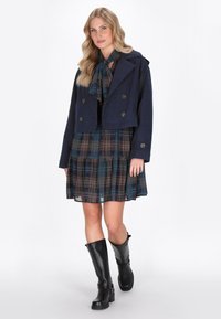 Marineblå cropped jakke med dobbeltknap lukning over en ternet kjole. Knæhøje sorte støvler fuldender outfittet. Glat stof og lagdelte detaljer.