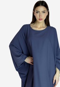 Vrouw met lang, donker haar in een losse, marineblauwe jurk met brede mouwen, staand met één hand op de heup tegen een witte achtergrond.