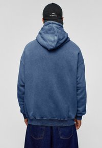 Blauwe hoodie gemaakt van katoen, met een ontspannen pasvorm en geribde manchetten. Een pet met de tekst "Sorry, ik ben geen DJ" wordt gedragen.