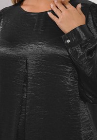 Top negro y brillante con una superficie texturizada, escote redondo y detalle de pliegue. Puños adornados con un acento de botón decorativo.