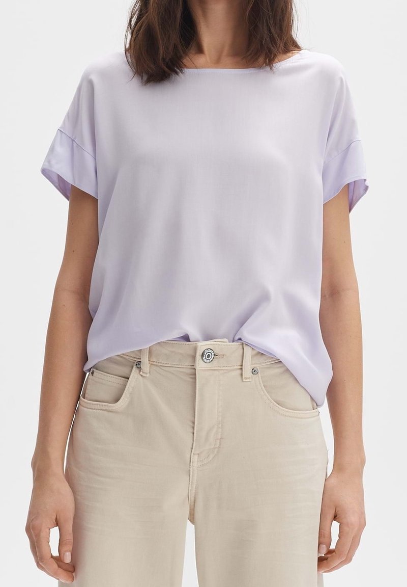 Femme portant un chemisier ample violet clair à manches courtes, rentré dans un pantalon beige taille haute avec des poches avant et une fermeture à boutons.