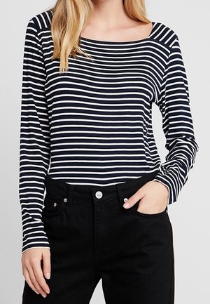 Camicia a maniche lunghe a righe in blu navy e bianco, con una scollatura ampia e una silhouette aderente. Abbinata a jeans neri a vita alta.