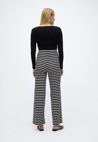 Γυναίκα που στέκεται με την πλάτη, φοράει μαύρο μακρυμάνικο τοπ, μαύρο-άσπρο παντελόνι με οριζόντιες ρίγες και μαύρα επίπεδα παπούτσια.