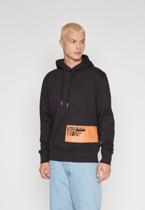Felpa nera con cappuccio, caratterizzata da una patch arancione con logo nella parte inferiore frontale. Tessuto morbido, vestibilità comoda e polsini a costine. Jeans blu chiaro.