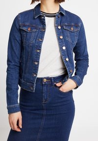 Mörkblå denimjacka med en figurnära design, knappstängning, två bröstfickor och kontrasterande sömmar, bärs över en grå skjorta.