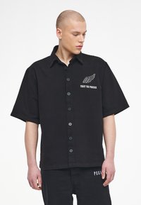 Black short-sleeve shirt made of cotton, featuring a button-up front, embroidered wing design, and "TRUST THE PROCESS" text on the chest.