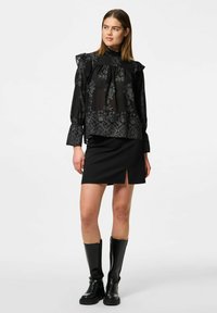 Blusa nera trasparente con motivo floreale, scollatura volants e maniche lunghe, abbinata a una mini gonna nera con apertura laterale. Stivali neri al ginocchio.
